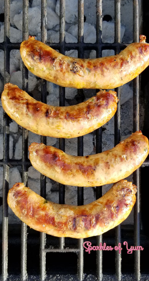 Four Homemade Jalapeno Cheddar Brats being grilled on a cast iron grate over charcoal.