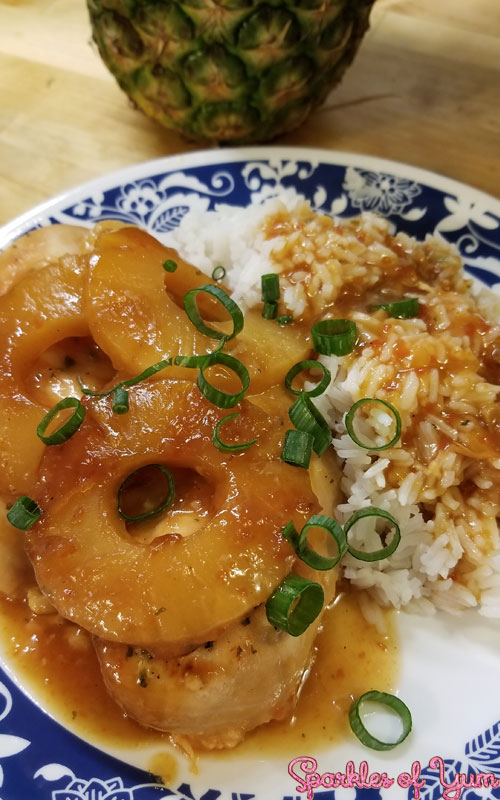 Chicken covered in a sticky sauce and pineapple rings on a white plate with blue detailing. There is white rice with the same sauce on the plate, and everything is garnished with sliced green onion.
