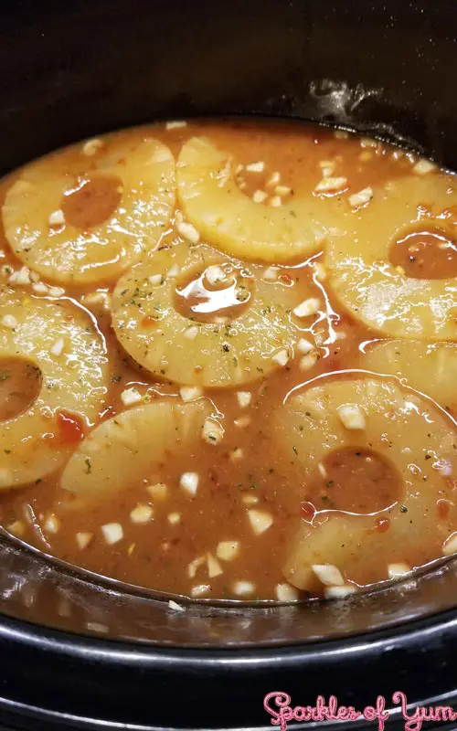 Pineapple rings and minced garlic floating in a crock pot filled with liquid.