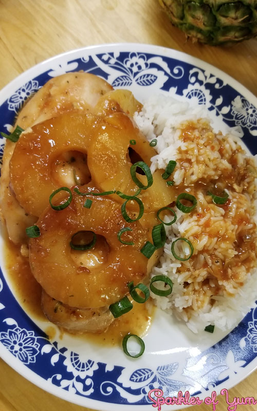Chicken and white rice covered in a light brown sauce. There is three pineapple rings on the chicken. Everything is arranged on a white plate with a blue pattern, and garnished with green onion.