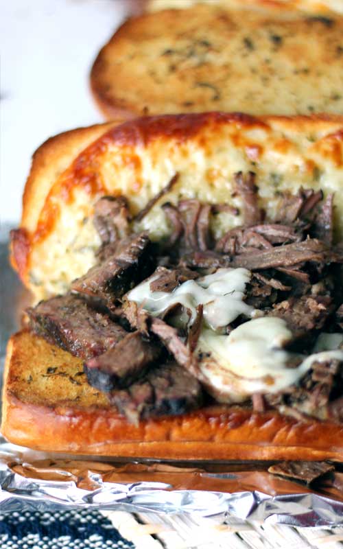 Smoked brisket topped with melted cheese. The meat is in a baguette that has been split open. and has bits of toasted cheese on the edges.
