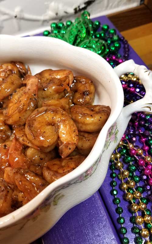 A white bowl containing New Orleans Barbeque Shrimp without the sauce. The bowl is resting on gold, purple, and green Mardi Gras beads.