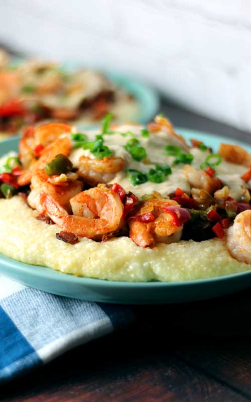 A blue plate with Shrimp and Grits on it. Green onion, cheese gravy, and red bell pepper cover the shrimp.