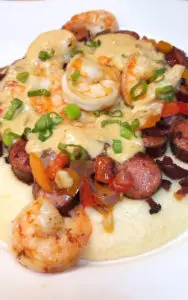Bring water, grits, and salt to a boil in a heavy saucepan with a lid. Stir in half-and-half and simmer until grits are thickened and tender, 15 to 20 minutes. Set aside and keep warm. Sprinkle shrimp with salt and cayenne pepper. Set aside in a bowl. Place andouille sausage slices in a large skillet over medium heat. Fry sausage until browned, 5 to 8 minutes. Remove skillet from heat. Cook bacon or Tasso in a large skillet over medium-high heat, turning occasionally, until evenly browned, about 10 minutes. Retain drippings in skillet. Transfer slices to paper towels. Cook and stir red, and yellow bell peppers, onion, and garlic in the bacon drippings until the onion is translucent, about 8 minutes. Remove to a plate. In the same skillet deglaze with white wine and cook shrimp 2-3 minutes. until the shrimp becomes pink. Remove to another plate. Melt butter in the same skillet over medium heat; to make a roux stirring for five minutes to incorporate.. Turn heat to low and cook, stirring constantly, until the mixture is light brown in color. Watch carefully, mixture burns easily. Pour in chicken broth, Louisiana Hot Sauce, Worcestershire Sauce, cooking and stirring until the sauce thickens, a couple minutes. If you feel like it's too thick you can add a little cream or milk. Just before serving, mix sharp Cheddar cheese into grits until melted and grits are creamy and light yellow. Plate grits on the bottom, andouille sausage and veggies over that with gravy and shrimp topping it all off and garnish with green onions.