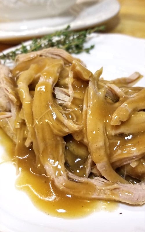 Close up of shredded Trisha Yearwood’s Crock Pot Pork Tenderloin covered in gravy, on a white plate. Fresh thyme sprigs are just out of focus in the background.