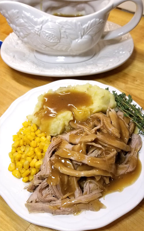 A white plate with corn, mashed potatoes and gravy, and shredded pork covered in gravy. In the background is a white gravy boat.