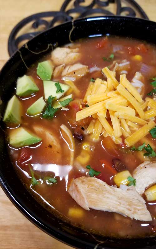 A black bowl filled with a reddish colored soup. In the soup cheddar cheese, shredded chicken, and diced avocado are all visible.