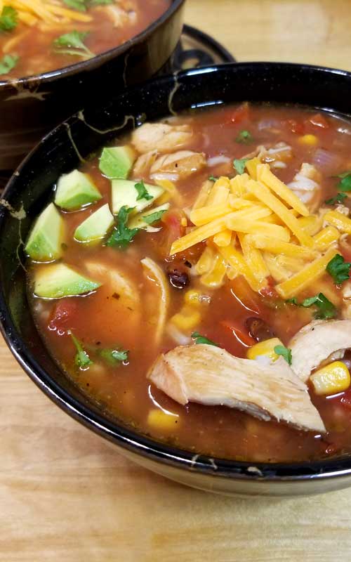 A black bowl filled with soup. The soup has cheddar cheese, diced avocado, and shredded chicken in it.