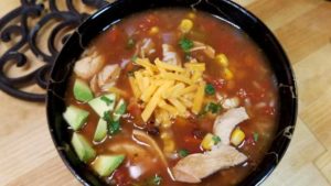 Top down view of a black bowl filled with soup. Shredded chicken, cheddar cheese, and diced avocado are visible within the soup.