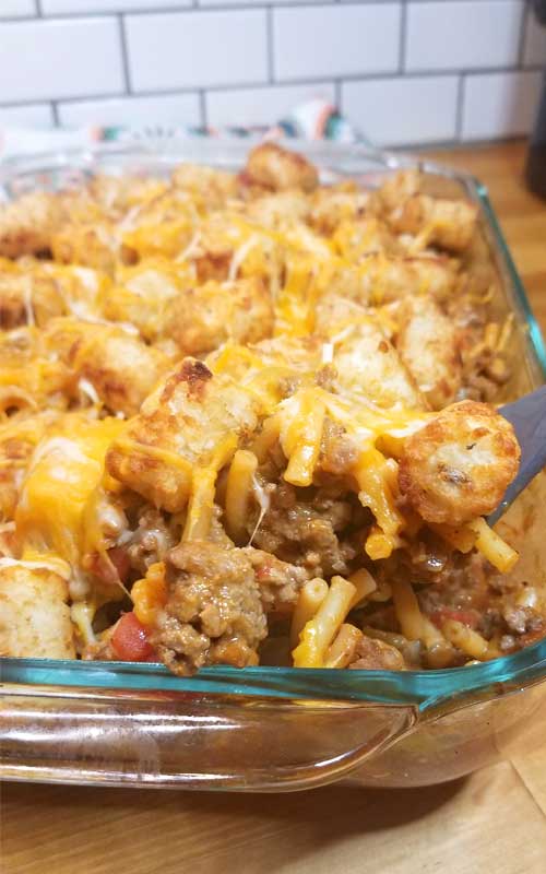 A spoon taking a scoop out of a Mac n Cheesy Cowboy Casserole that is in a glass baking dish.