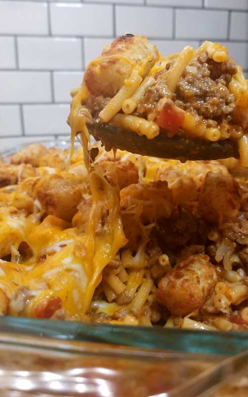 A finished Mac n Cheesy Cowboy Casserole in a glass baking dish. A spoon is taking a scoop out of the casserole.