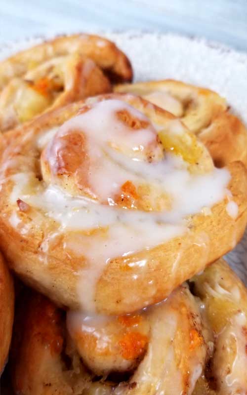 A pile of Orange Pineapple Sweet Rolls on a white platter. The rolls are covered in white icing, and pieces of orange can be seen in the bread.