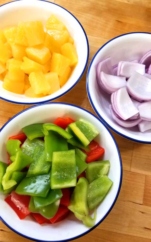 Three white bowls. One is filled with pineapple chunks, one with pieces of red onion, and the last with pieces of red and green bell peppers.