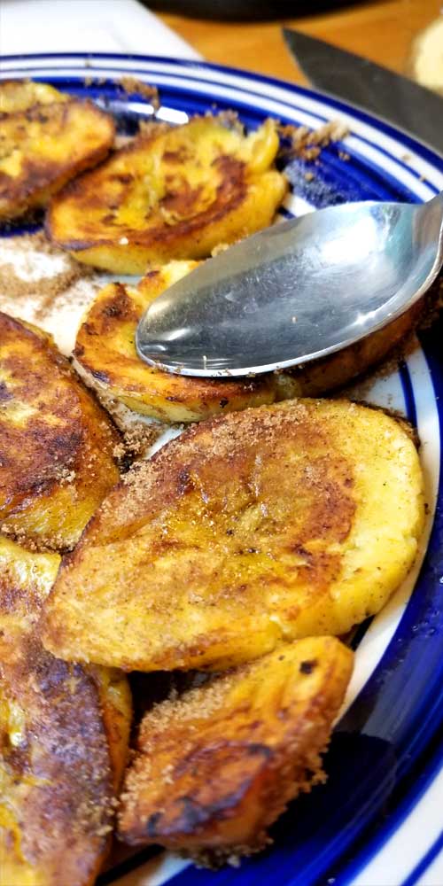 Sweet Fried Plantains on a white plate with blue trim. There is a spoon smashing one of the plantains.