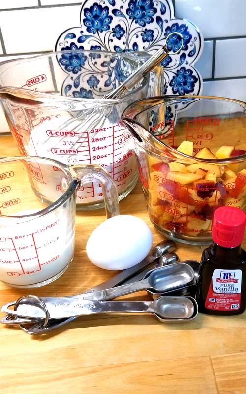 Three clear glass measuring cups; one with milk, one with flour, and the last with diced peaches. There is also one egg, a set of metal measuring spoons, and a bottle of vanilla extract.