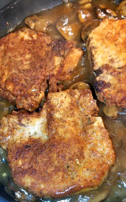 Three fried pork chops in a non-stick skillet, being cooked with muchrooms and gravy.