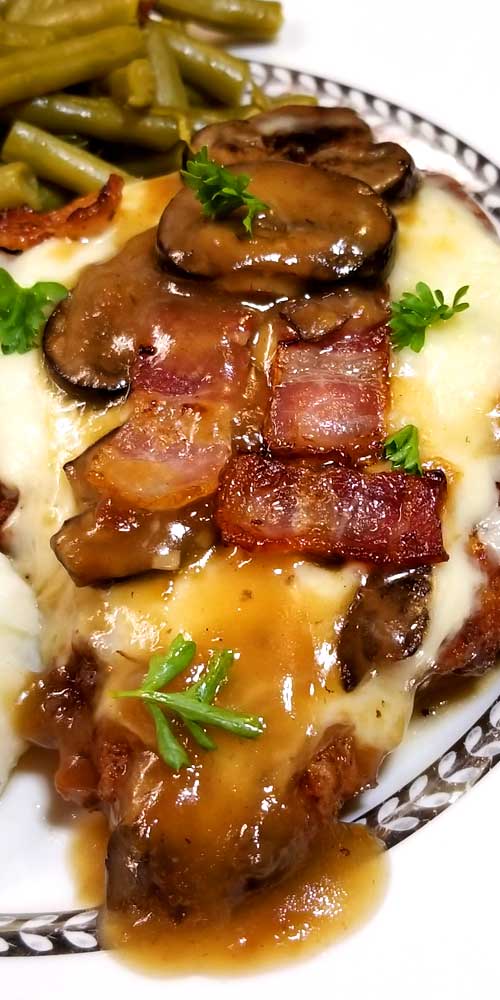 A cooked pork chop covered with Fontina cheese, bacon, mushrooms, a mushroom gravy, and parsley for garnish.