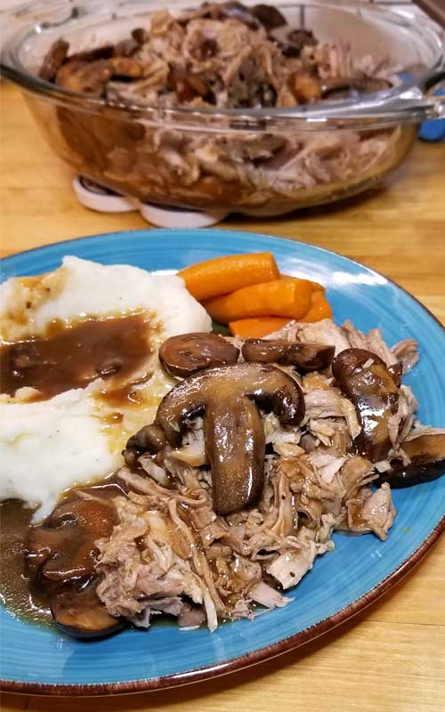 Shredded pork loin and mushrooms covered in a mushroom sauce, with carrots and mashed potatoes and gravy on a blue plate. In the background is a clear glass bowl with more shredded pork loin.