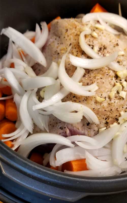 A seasoned pork loin covered in onions sitting on top of chunks of carrot, in a black non-stick cooking pot.