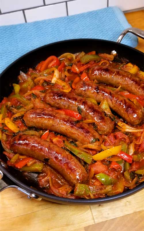 A skillet filled with five Italian sausages plus red, yellow, and green peppers. Part of the skillet is resting on a light blue towel, against a tile backsplash.
