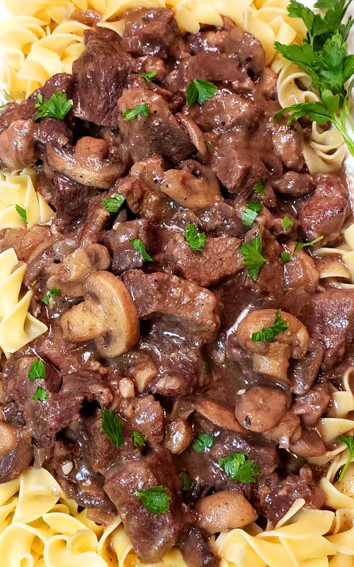 Top-down view of Garlic Butter Beef Tips and Mushroom Gravy over egg noodles. A small bunch of parsley is in the top right corner.