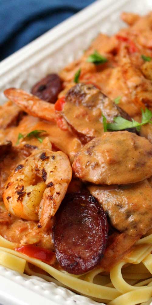 A white serving platter with shrimp and sliced sausage covering pasta. The shrimp and sausage are covered in a cream sauce.