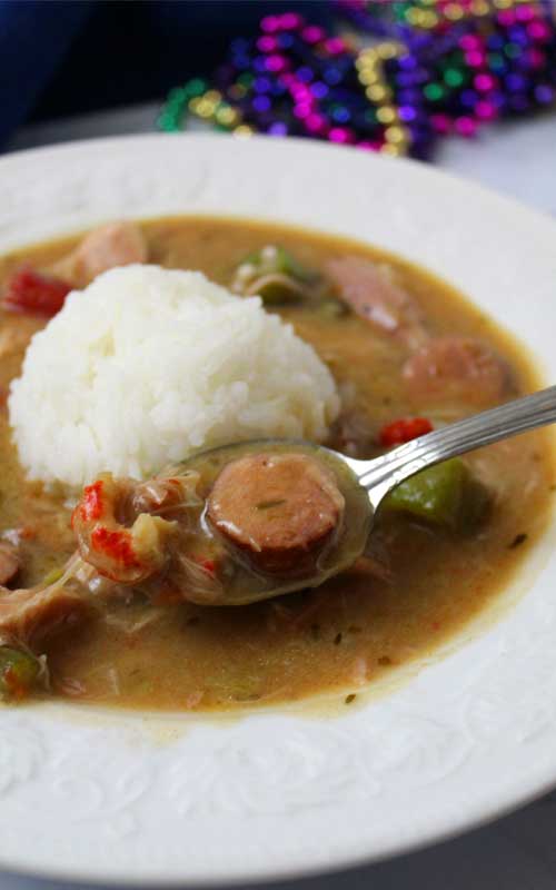 Authentic New Orleans Cajun Gumbo and white rice in a white bowl. There is a spoon that has scooped out a piece of sausage and crawfish tails.