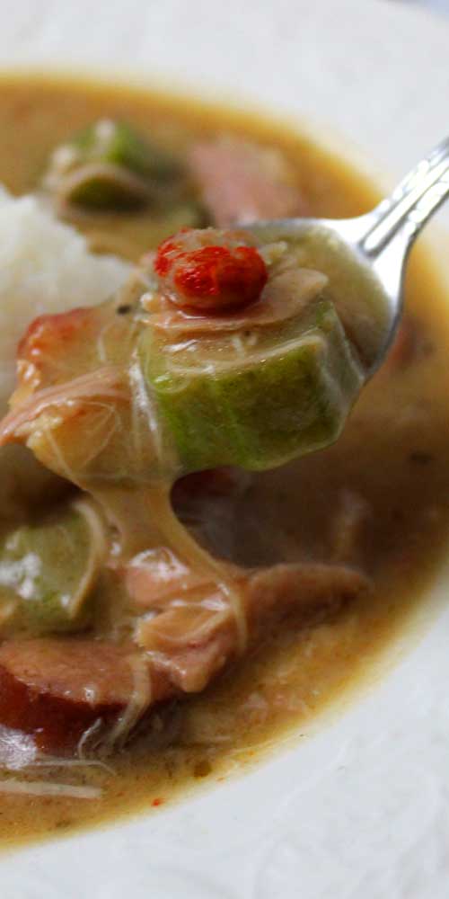 Close up view of a spoon that has scooped out okra and crawfish from a white bowl filled with Authentic New Orleans Cajun Gumbo.