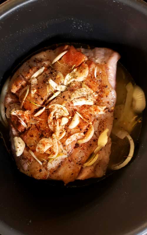 Uncooked pork roast sitting in crock pot. Roast is covered with garlic and seasonings.