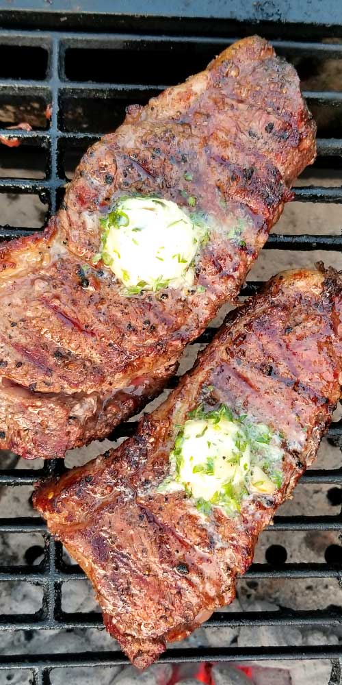 Two Grilled Rib-Eye Steaks with Roasted Garlic Herb Butter on a cast iron grate.