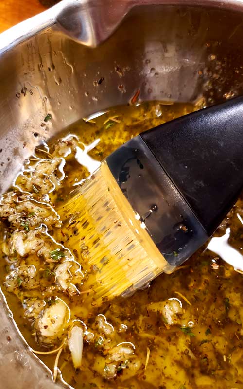 Close up view of olive oil, garlic, and herbs being stirred in a metal bowl with a basting brush.