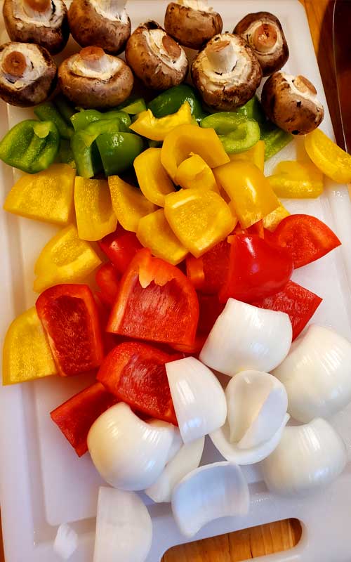 Pieces of green, yellow, and red bell pepper; along with pieces of onion, and baby bella mushrooms resting on a white cutting board.