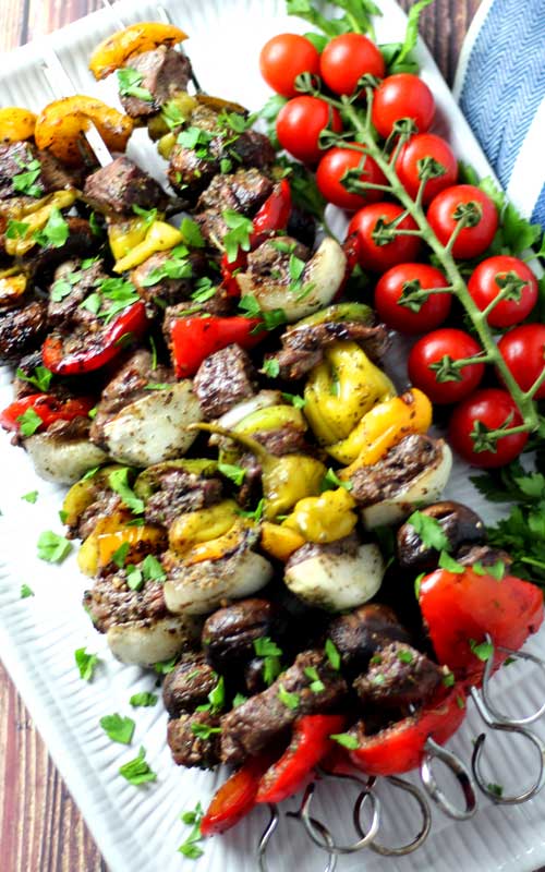 An over head view of steak kabobs with various vegetables resting on a white platter. A vine of cherry tomatoes rests on the right edge of the platter.