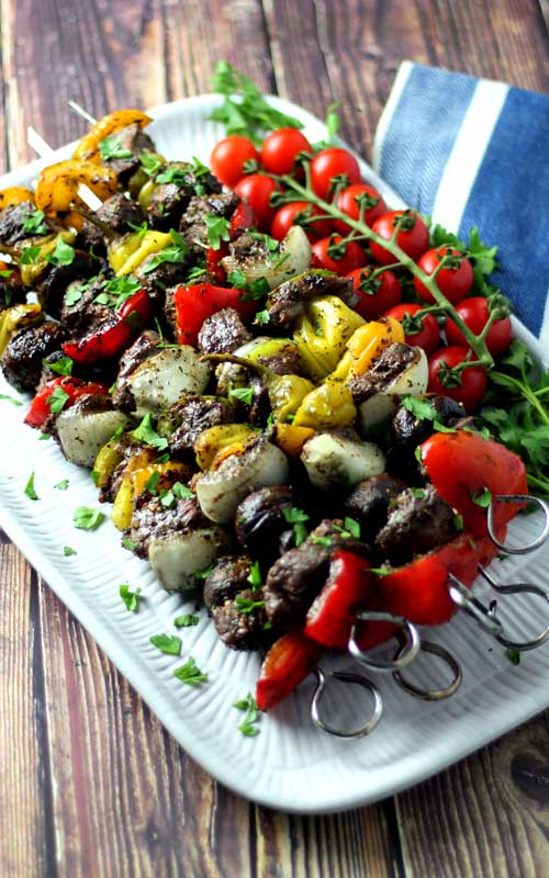 Grilled vegetables and pieces of steak on metal skewers. The food is arranged on a white platter.