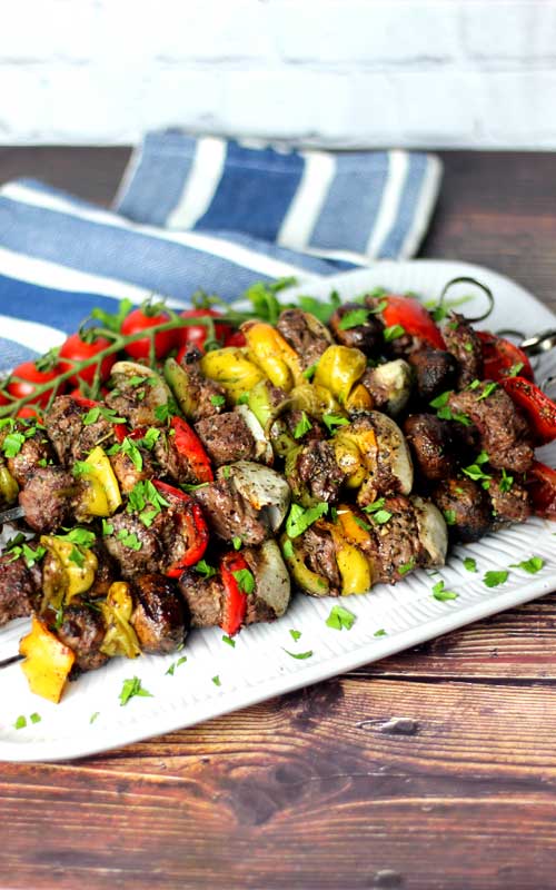 A white platter with grilled kabobs on it. The kabobs are made of steak, onion, red bell pepper, pepperoncini, and mushrooms. A vine of red cherry tomatoes and a blue towel can be seen in the background.