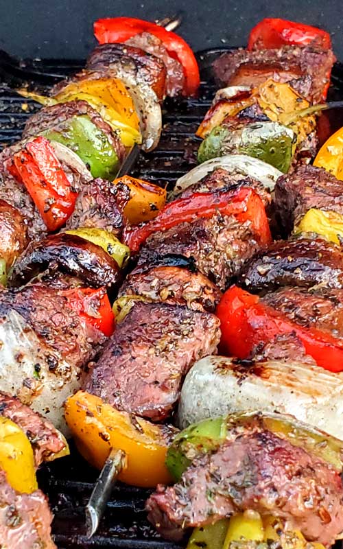 Steak, onion, and bell peppers pierced with a metal skewer. The food is being grilled.