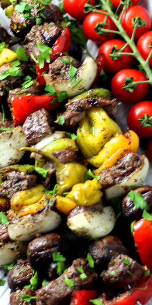 Very close up view of grilled steak, pepperoncini, mushrooms, bell pepper, and onion after being grilled. Res cherry tomatoes on the vine are visible in the top right corner of the image.