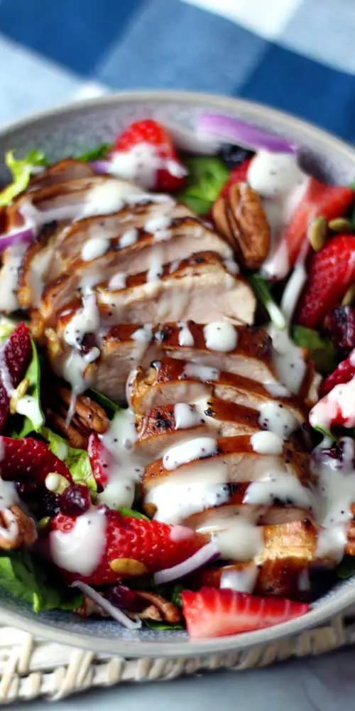 This Summer Strawberry Spinach Salad with Chicken is so tasty and refreshing for a hot day. It's simple, healthy, and incredibly delicious! A white bowl filled with a salad made of spinach, sliced strawberries, and grilled chicken. A white dressing is on the salad. A blue and white checkered towel is in the background.