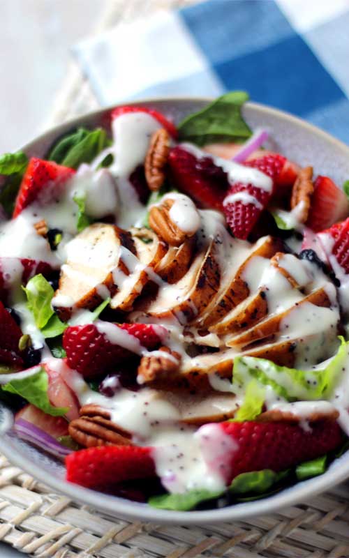 Grilled chicken, strawberries, lettuce, and pecans are arranged in a white bowl. A white colored dressing is on the salad.