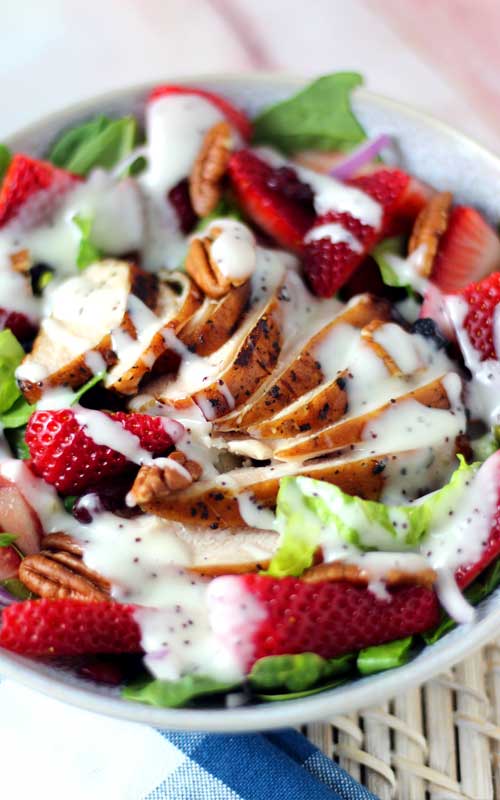 A white colored salad dressing tops grilled chicken, lettuce, pecans, and sloced strawberries. The salad is arranged in a white bowl.