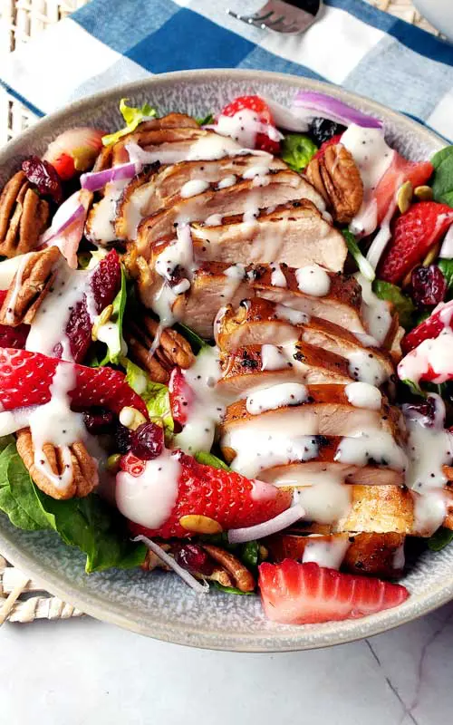An overhead view of a prepared salad. The salad is made with lettuce, pecans, strawberries, and grilled chicken, The dish is topped with a white colored salad dressing.