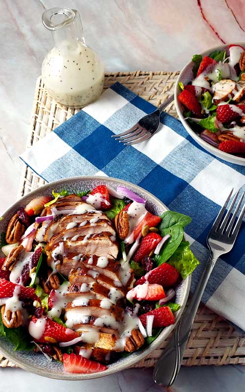 Two white bowls are filled with salad. The salad is made from grilled chicken, sliced strawberries, pecans, and lettuce. There is a fork next to each bowl. In the top left of the image is a small pourer filled with dressing.