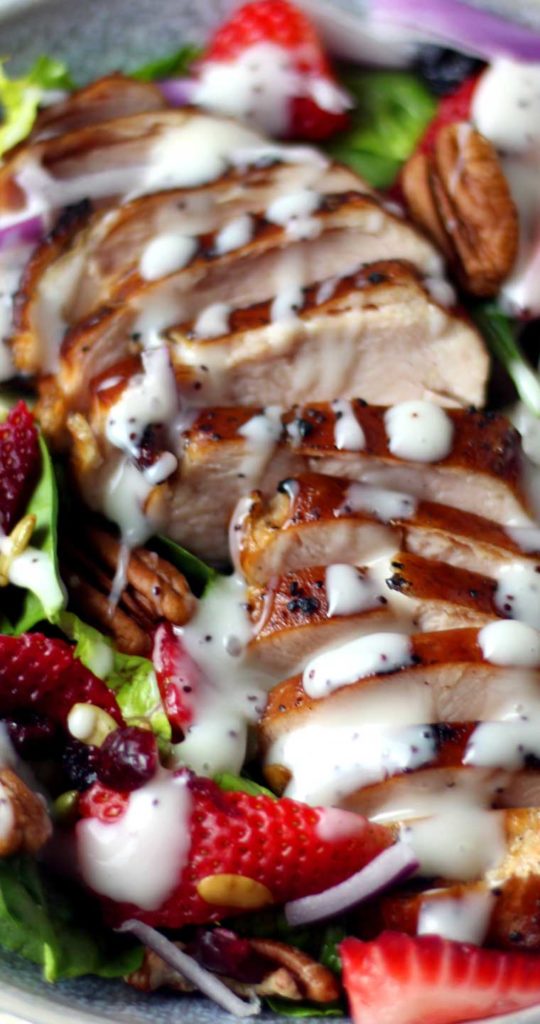 A close up view of sliced grilled chicken covered with a drizzle of white salad dressing. Strawberries, pecans, and lettuce are visible to the sides of the chicken.