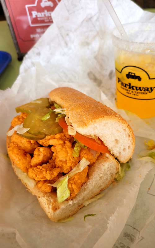 A Parkway Bakery Shrimp Po'Boy with lettuce, tomato, and pickles on white butcher paper. In the background is a drink in a Parkway Bakery cup.