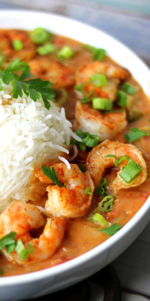 A white bowl filled with shrimp etouffee. The shrimp are in an orange colored, vegetable gravy. There is a scoop of white rice in the bowl, and the dish is garnished with parsley and green onions.