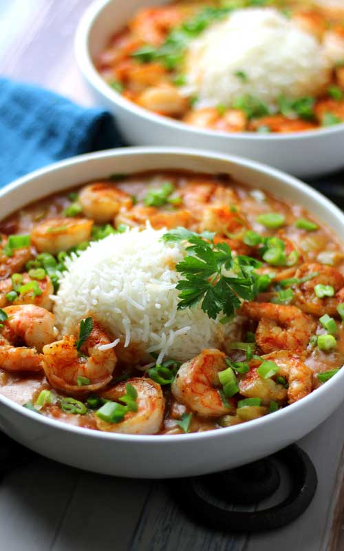 Two shallow white bowls filled with shrimp etouffee. Many shrimp are sitting atop a tomato based gravy. In the center of each bowl is a mound of white rice. Each bowl has parsley and green onion for garnish.