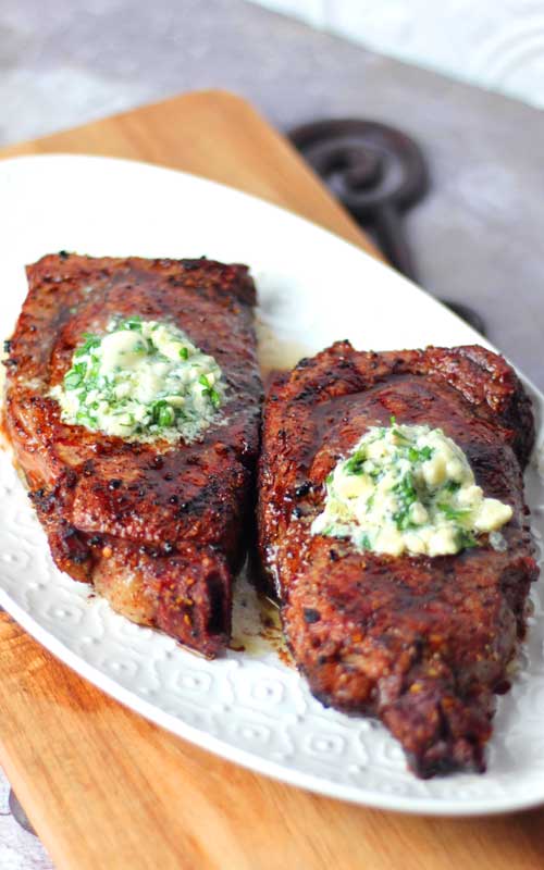 Two Grilled Ribeye Steaks topped with Gorgonzola Herb Butter resting on a white platter.