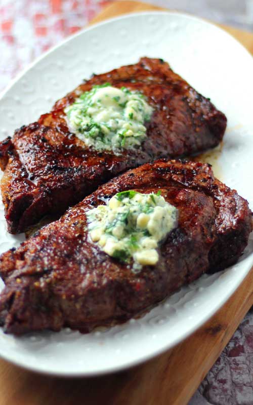 Two Grilled Ribeye Steaks topped with Gorgonzola Herb Butter resting on a white platter.