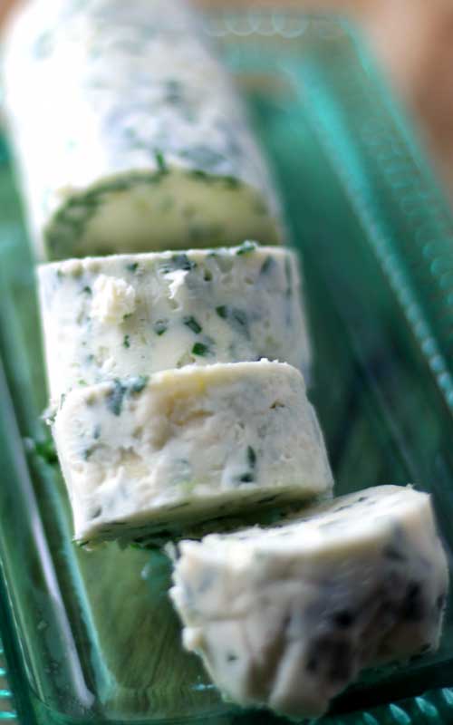 A round stick of Gorgonzola Herb Butter that has had three pieces cut out of it. The butter is resting on a green, glass dish.