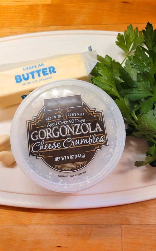 A stick of butter, fresh parsley, garlic cloves, and a container of gorgonzola cheese sitting on top of a white cutting board.